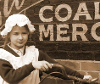 Black and white image of a young child dressed up in Victorian clothing wearing a white cloth hat.