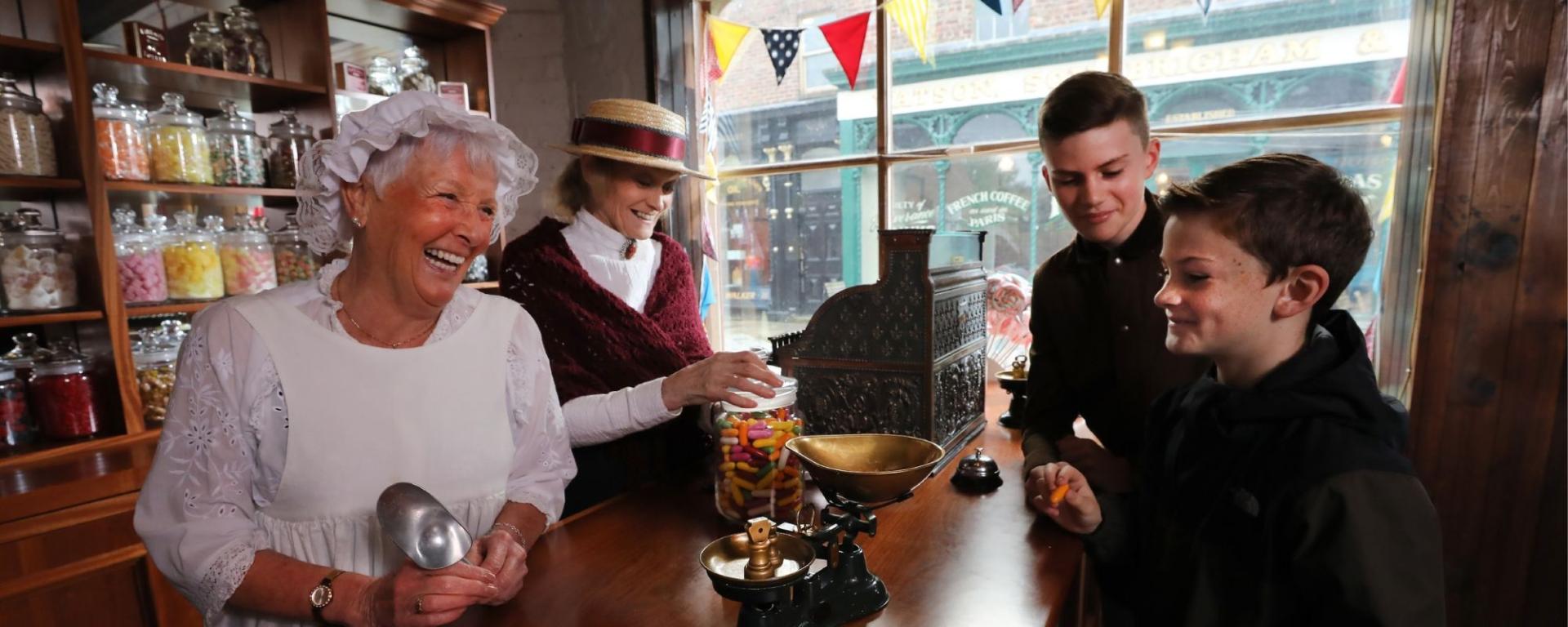 Young Boys Being Served Old Fashioned Sweets By Two Ladies In Victorian Costume, In The Sweet Shop Young Boys Being Served Old Fashioned Sweets By Two Ladies In Victorian Costume, In The Sweet Shop